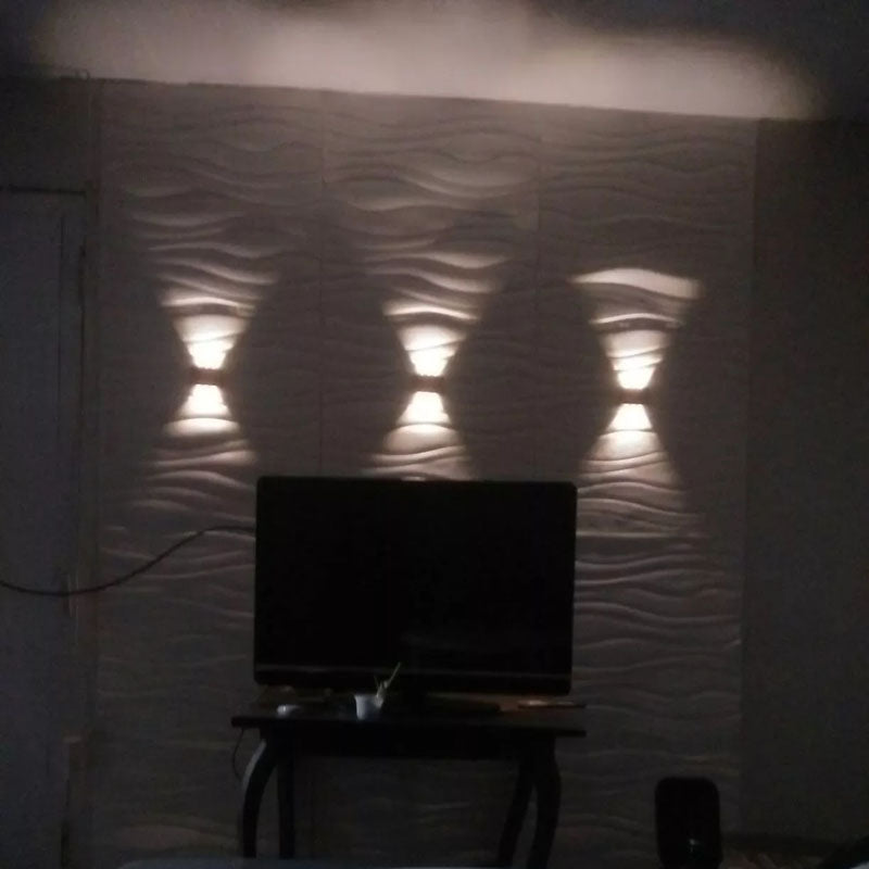 A solar wall lamp installed on a garden wall, providing illumination to the area with its LED beads lighting. The wall lamp is mounted on a sturdy, weatherproof structure with a pattern of panels and a visible solar panel on the ground.