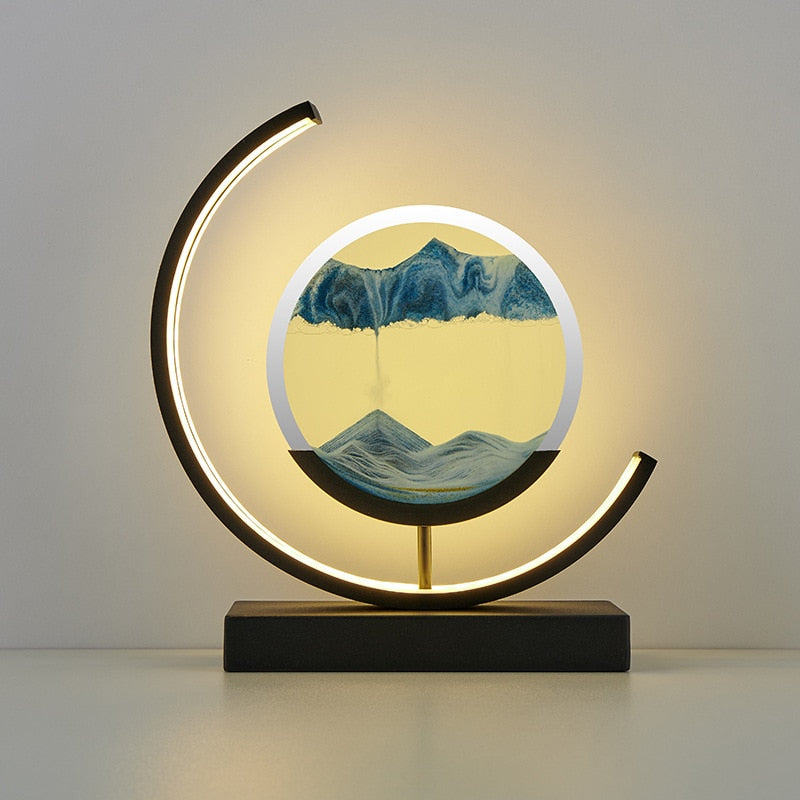 A unique decorative LED desk lamp with a 3D sand art design that creates a serene landscape, placed on a bookshelf in a home environment.